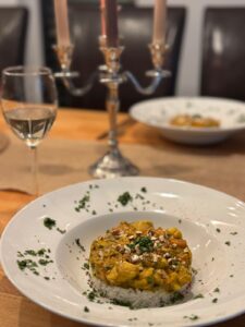 Ein Teller Curry mit Reis ist mit gehackten Nüssen und Kräutern garniert. Im Hintergrund stehen ein Glas Weißwein und ein brennender Kerzenleuchter auf einem Holztisch. Ein weiterer ähnlicher Teller ist teilweise sichtbar.