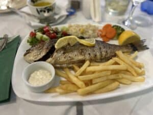 Ein Tellergericht mit einem ganzen gegrillten Fisch, garniert mit Zitronenscheiben, dazu knusprige Pommes Frites, Remoulade und ein frischer Salat mit Kirschtomaten, Gurke und Kopfsalat. Im Hintergrund stehen eine Zitronenscheibe.