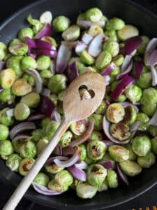 Eine Pfanne mit halbiertem Rosenkohl und gehackten roten Zwiebeln. Ein Holzlöffel mit herzförmiger Aussparung liegt auf dem Gemüse. Der Rosenkohl ist leicht gebräunt, was darauf schließen lässt, dass er angebraten wird.