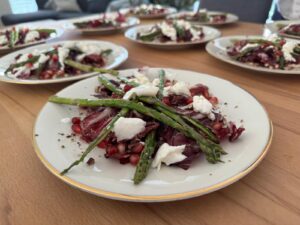 Mehrere Teller Salat mit gegrilltem Spargel, Radicchio, Ziegenkäse und Granatapfelkernen sind auf einem Holztisch angerichtet. Der Fokus liegt auf dem jeweils nächstgelegenen Teller, wodurch die frischen Zutaten hervorgehoben werden.