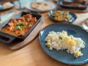 Eine Nahaufnahme einer Mahlzeit: Ein blauer Teller mit weißem Reis, garniert mit Kräutern. Daneben steht ein Tablett mit Curry in einer schwarzen Schale. Im Hintergrund sind ein kleiner Teller mit gemischtem Gemüse und anderen Gerichten leicht verschwommen.